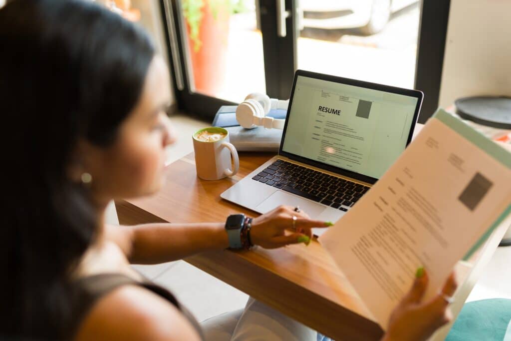 Young woman in a coffee shop applying for a job on a laptop, holding her resume.